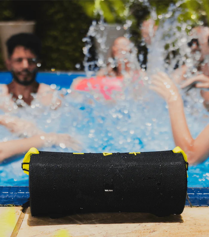 Caixa de som Bluetooth WAAW ao lado de uma piscina, com pessoas se divertindo ao fundo.