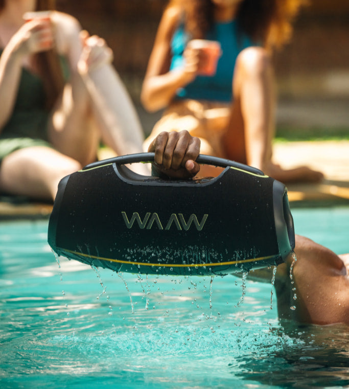 Pessoa segurando uma caixa de som bluetooth WAAW à beira da piscina, com água caindo do produto.