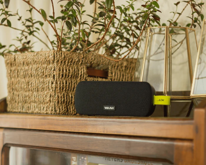 Caixa de som Bluetooth da marca WAAW sobre uma superfície de madeira, com plantas ao fundo.