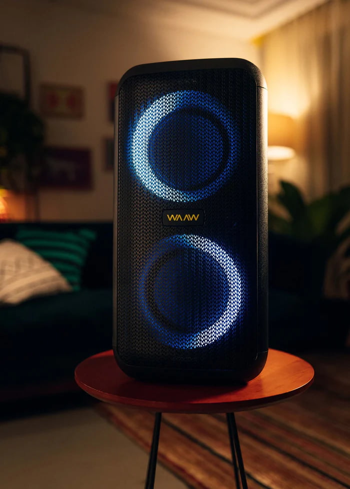 Caixa de som Bluetooth da marca WAAW com luzes azuis acesas, sobre uma mesa em um ambiente interno.