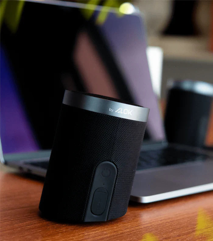 Caixa de som Bluetooth da marca WAAW, com logo "by ALOK", ao lado de laptop em uma mesa de madeira.